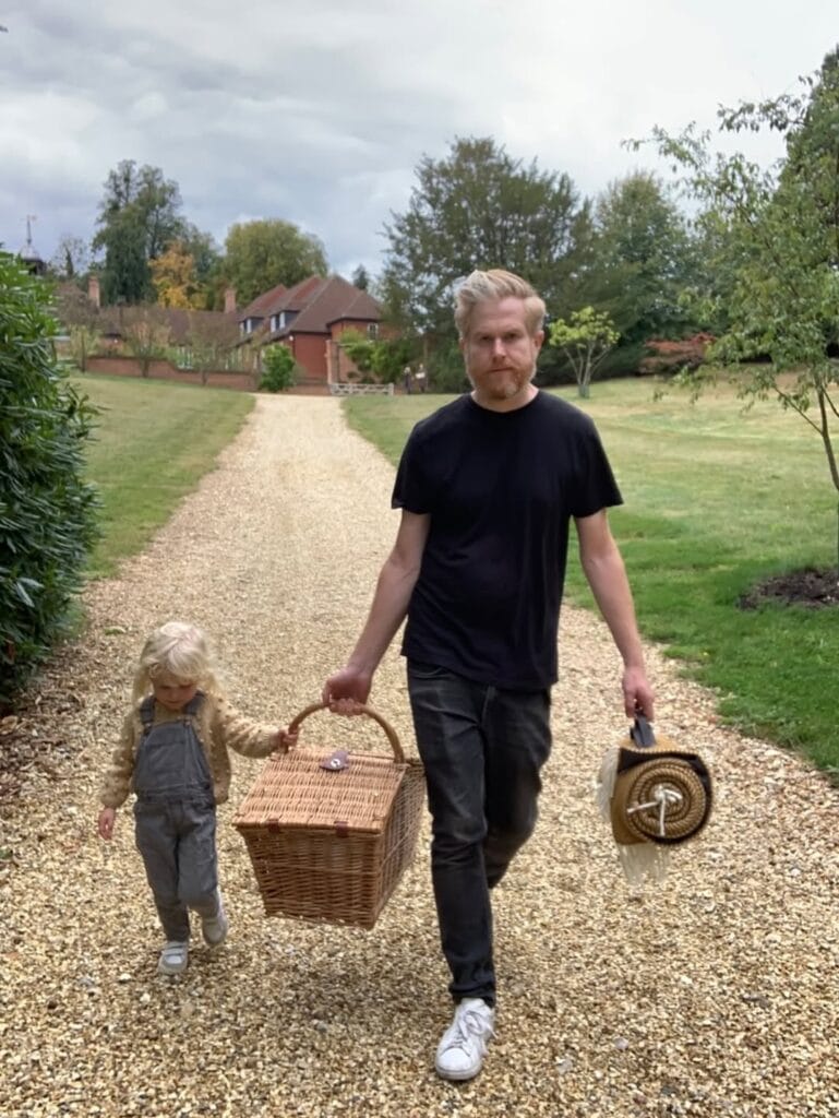 Paul Ferney and a young child in denim overalls walking down a gravel path together, sharing the handle of a wicker picnic basket, with a country house and green lawn in the background