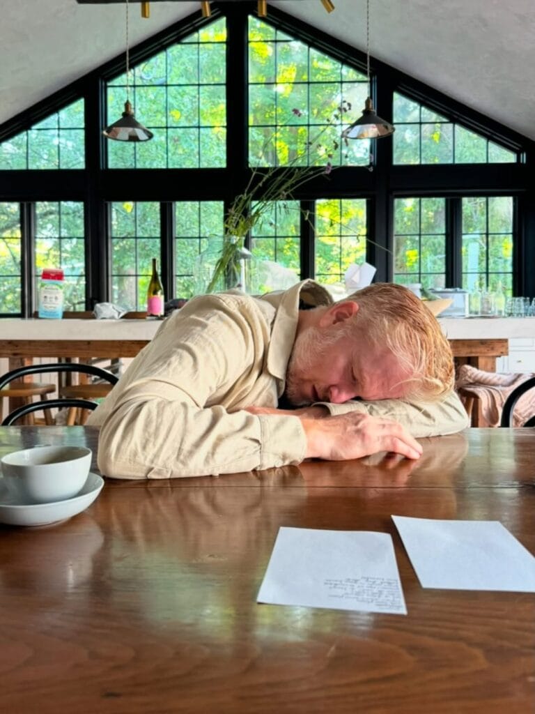 Paul Ferney resting his head on his arms at a wooden table in a bright room with large black-framed windows, a bowl and handwritten papers nearby