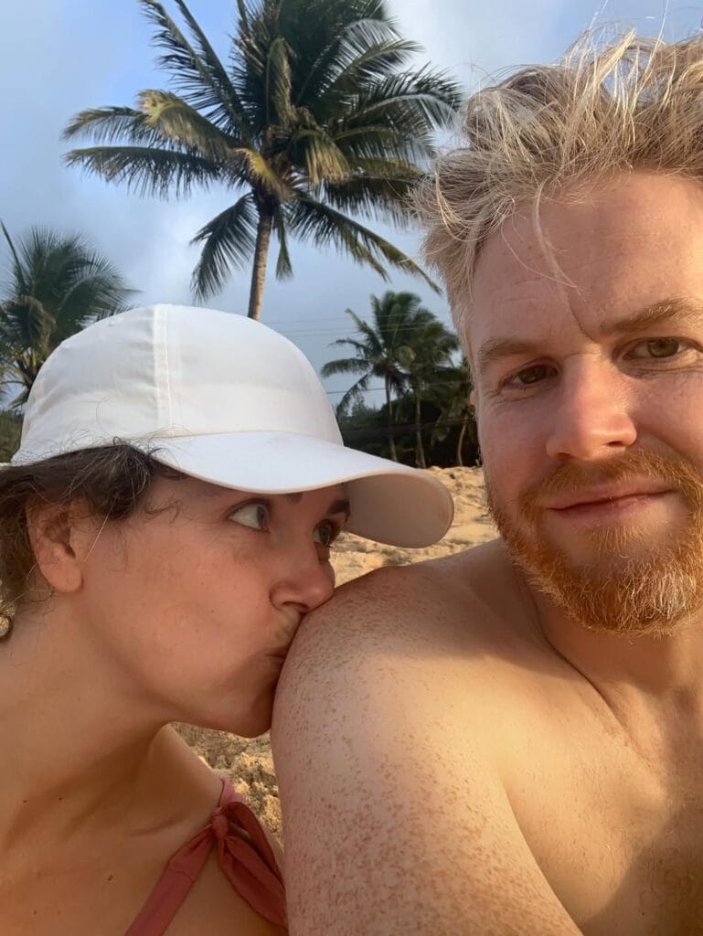 Jordan Ferney and a woman in a white baseball cap on a sandy beach with palm trees, she kisses his shoulder