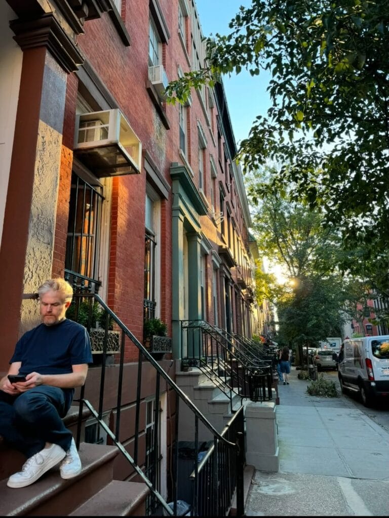 Paul Ferney sitting on brownstone stoop steps looking at his phone, tree-lined New York City sidewalk at golden hour