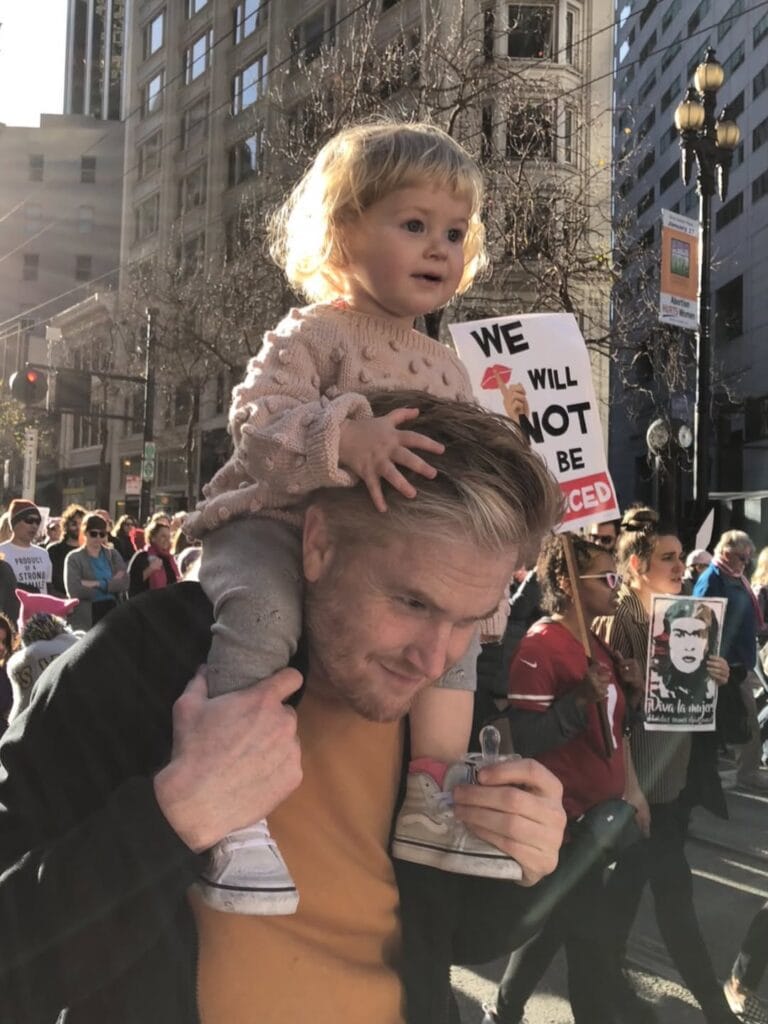Paul Ferney carrying a toddler on his shoulders at a street march, protest signs visible in the crowd behind them