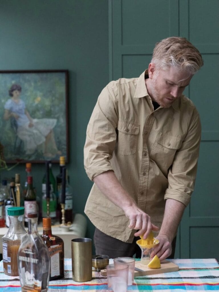 Jordan Ferney squeezing citrus into a juicer at a table set with cocktail bottles, a shaker, and a jigger, against dark green paneled walls
