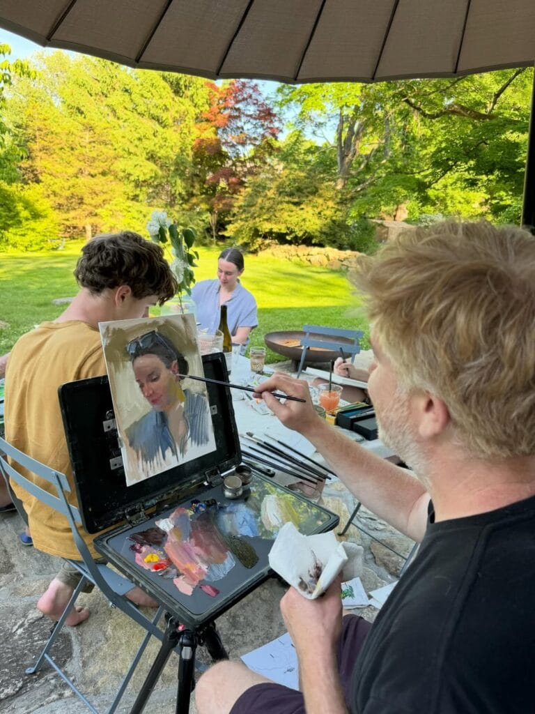 Paul Ferney painting a portrait on an easel at an outdoor patio gathering, lush garden in the background