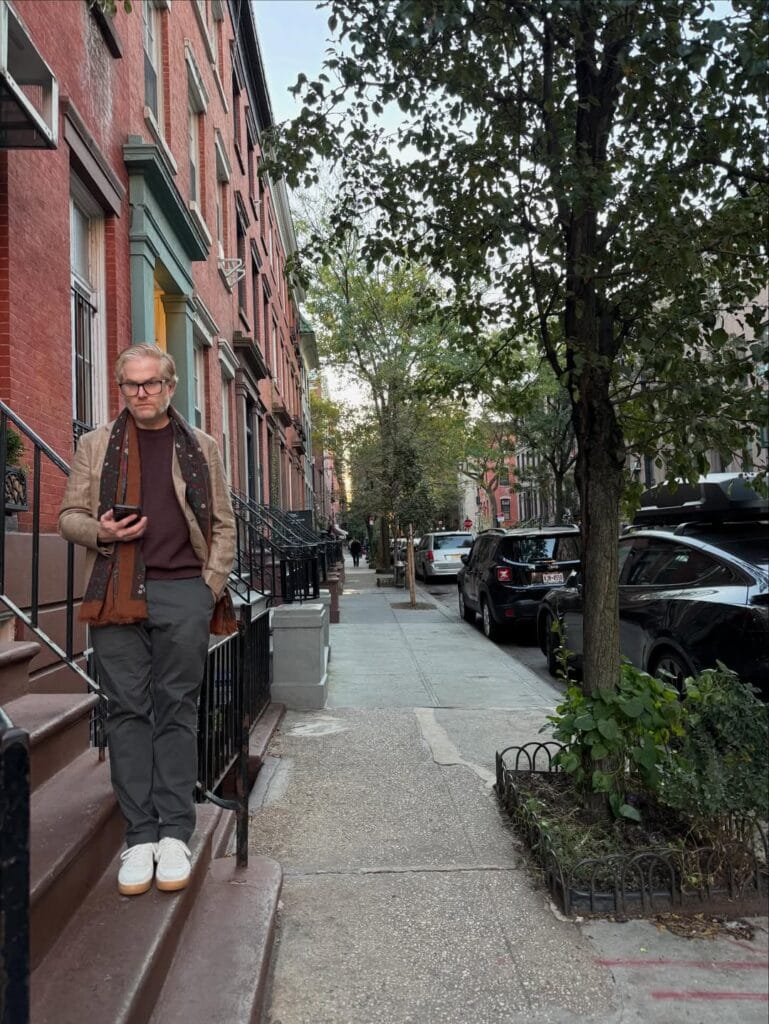 Paul Ferney walking down brownstone steps on a tree-lined city sidewalk, holding a phone, wearing a tan blazer and dark scarf
