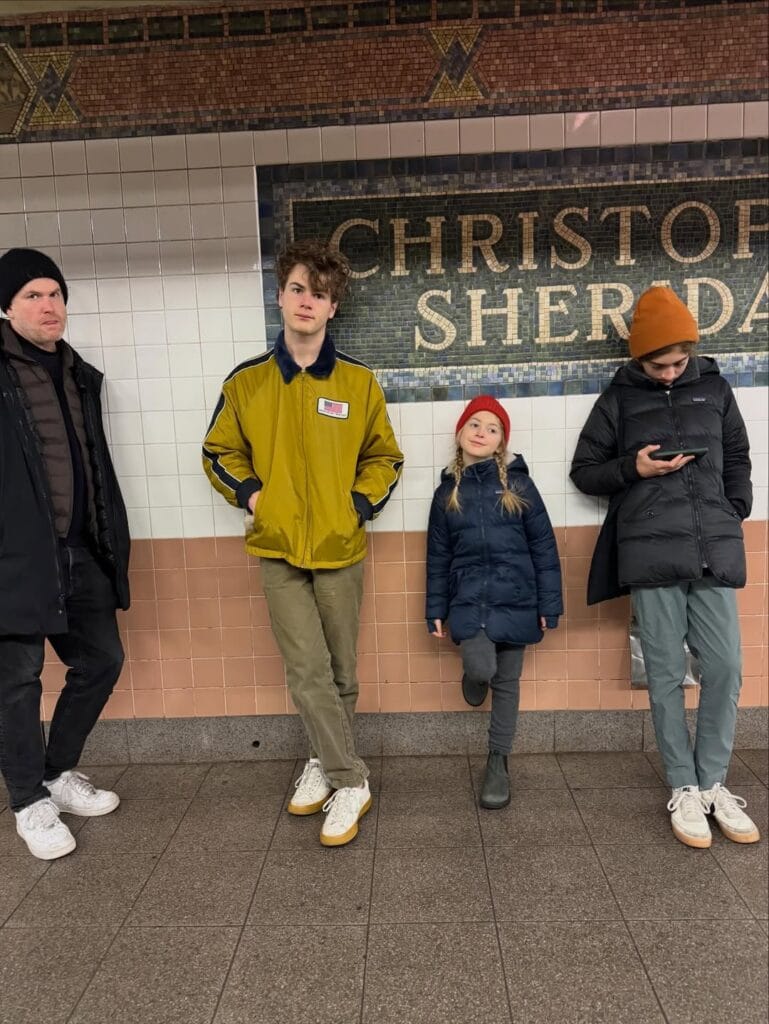 Four people standing on a New York City subway platform in front of the Christopher Street Sheridan Square station mosaic sign